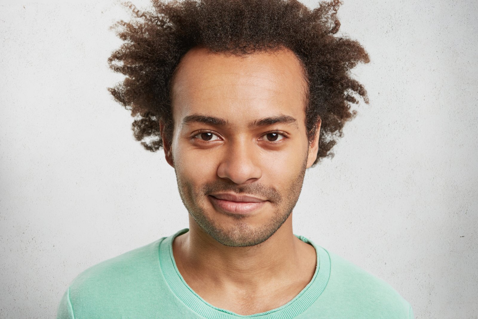 Headshot of stylish hipster guy with bushy hairstyle, stubble, shining eyes, poses in white studio, has pleased expression as hears pleasant comments from clients. Good looking stylish male student